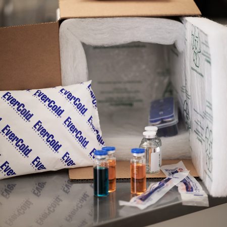 A open white insulated foam shipping box packed with medical diagnostic vials, needles, and a blue EverCold gel refrigerant pack, demonstrating a secure cold chain for clinical and pharmaceutical distribution. Representing EverCold gel ice packs for shipping