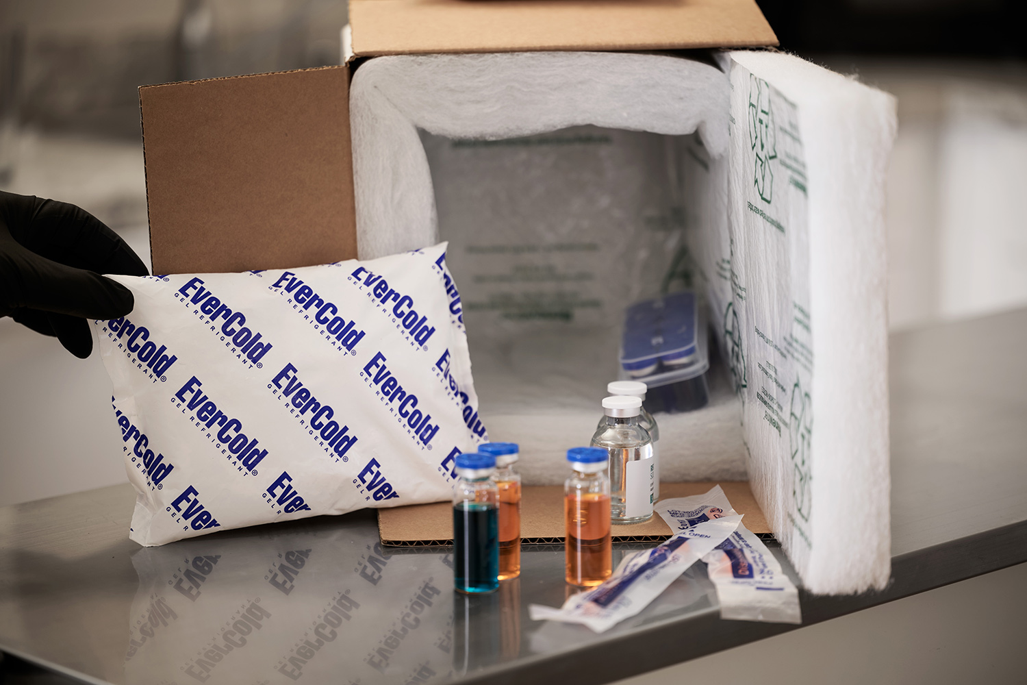 A open white insulated foam shipping box packed with medical diagnostic vials, needles, and a blue EverCold gel refrigerant pack, demonstrating a secure cold chain for clinical and pharmaceutical distribution. Representing EverCold gel ice packs for shipping