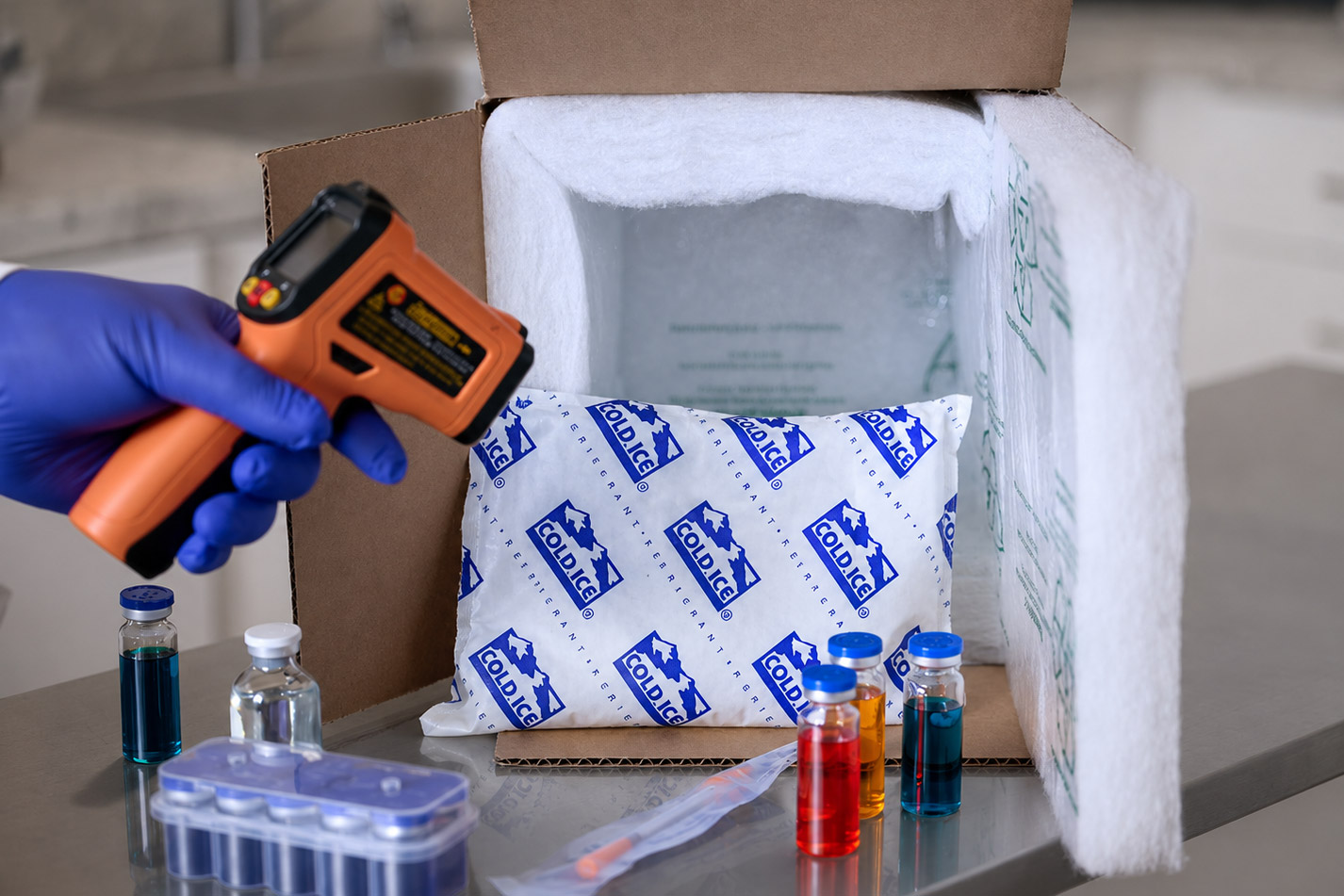 Gloved hand holding an orange infrared thermometer aiming at Cold Ice refrigerant gel packs inside an insulated cardboard box packed with glass sample vials.