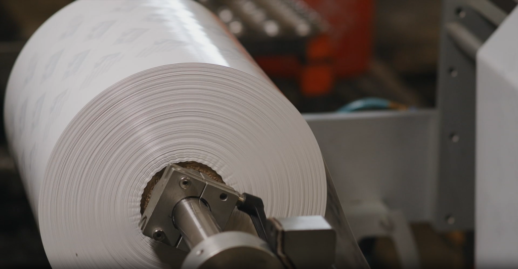 A close-up of a large roll of white plastic or paper lining material mounted on an industrial spooling machine roll inside a manufacturing or packaging facility.